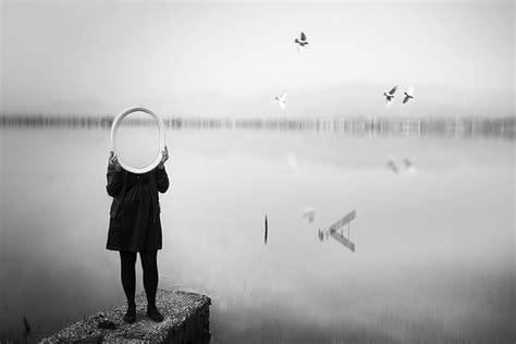 Person holding round mirror in front of face, standing on a pier by a calm lake. Four birds fly above. Black and white, dreamy mood.