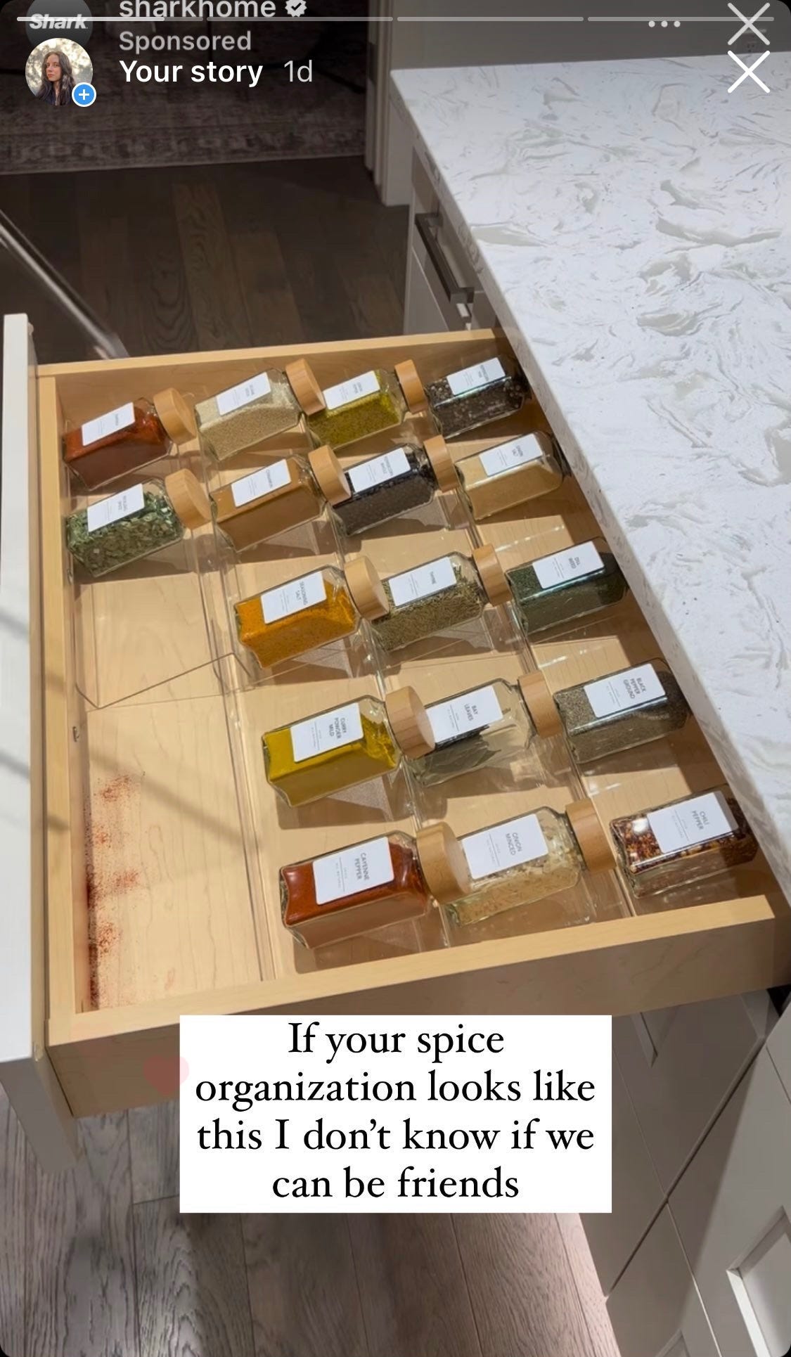 A screenshot of an ad on Instagram for Shark Home, showing a white woman vacuuming her spice drawer. A transparent plastic shelf system in the drawer keeps the bottles of spices displayed with their label face up. The drawer is about 50% full, 2-3 inches between each spice bottle. I reposted the screenshot, adding “If your spice organization looks like this I don't know if we can be friends.” 