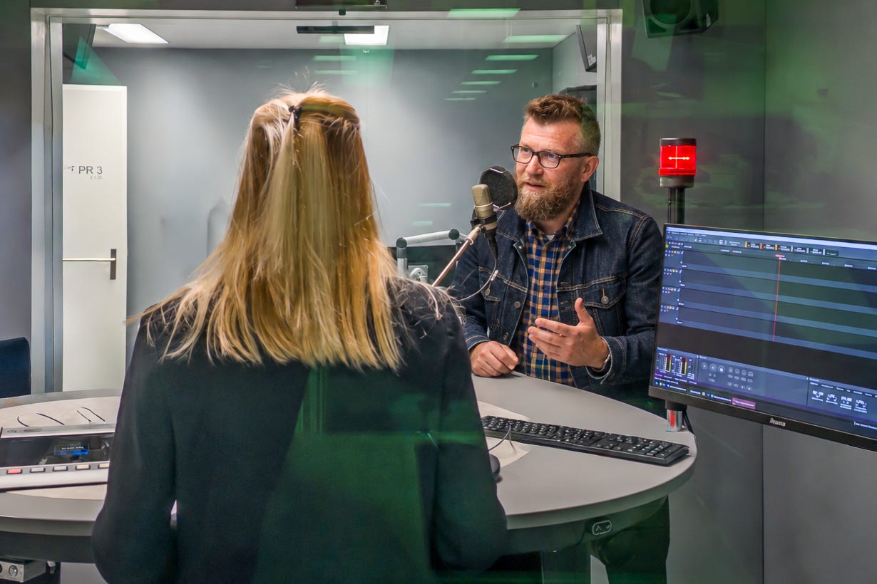 Interview im Radiostudio: Jürg Vollmer mit Bart und Brille steht einer blonden Rediojournalistin gegenüber und spricht gestikulierend in ein Mikrofon. Rechts ist die Audiotechnik zu sehen. Darunter der Schriftzug «Grüezi Servus & Hallo».
