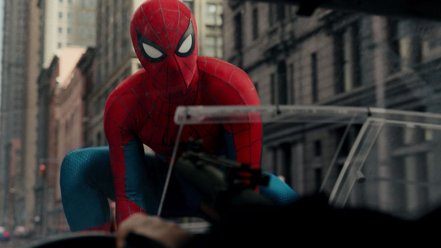 Spider-Man squats on the hood of a truck, looking through the window at its driver