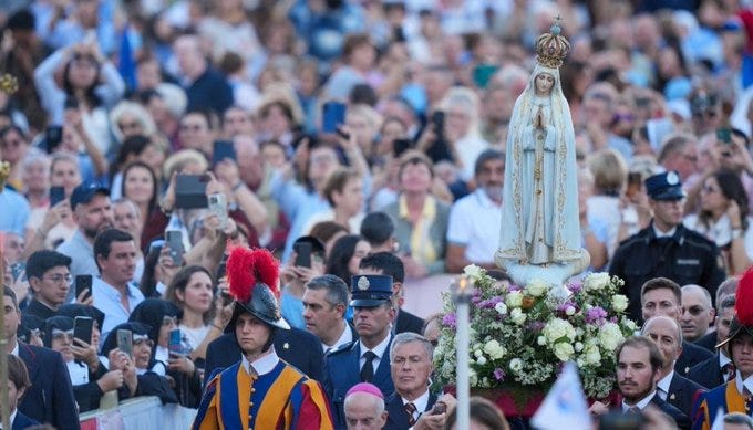 A large crowd gathers outdoors during a religious procession, with participants in formal attire including red-plumed hats, uniforms, and clerical robes carrying a white statue of Our Lady of Fatima adorned with a crown and surrounded by flowers on a pedestal. People hold phones to take photos, and the scene includes security personnel and diverse attendees in casual clothing under a clear sky.