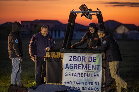 Înainte ca baloanele să urce, totul începe jos, pe iarbă, în frigul subțire al dimineții sau în lumina care abia se stinge peste câmp. Oamenii își ocupă locurile, verifică arzătoarele, întind pânza, ridică nacela, privesc cerul și, mai ales, ascultă vântul. În jur, mașinile stau cu farurile aprinse.