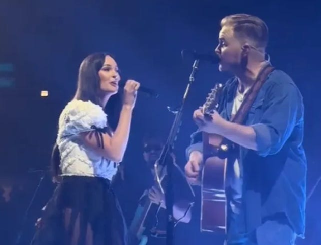 Country artists Kacey Musgraves and Zach Bryan perform on stage together, with Kacey holding a microphone and Zach holding a guitar.