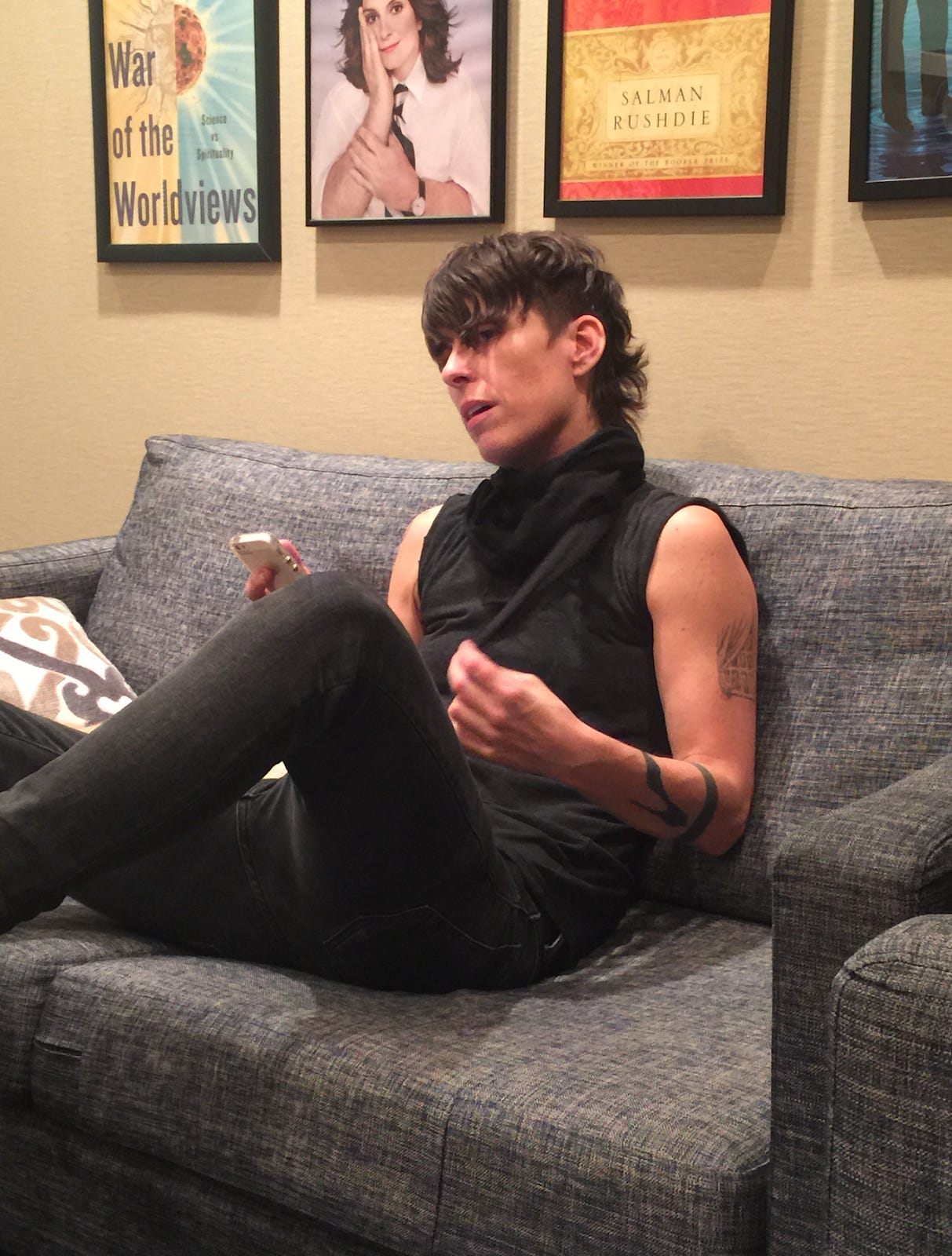 Andrea Gibson sits on a couch in a backstage area. They are speaking to someone out of frame. They wear black jeans, a black muscle tank, and a black scarf.