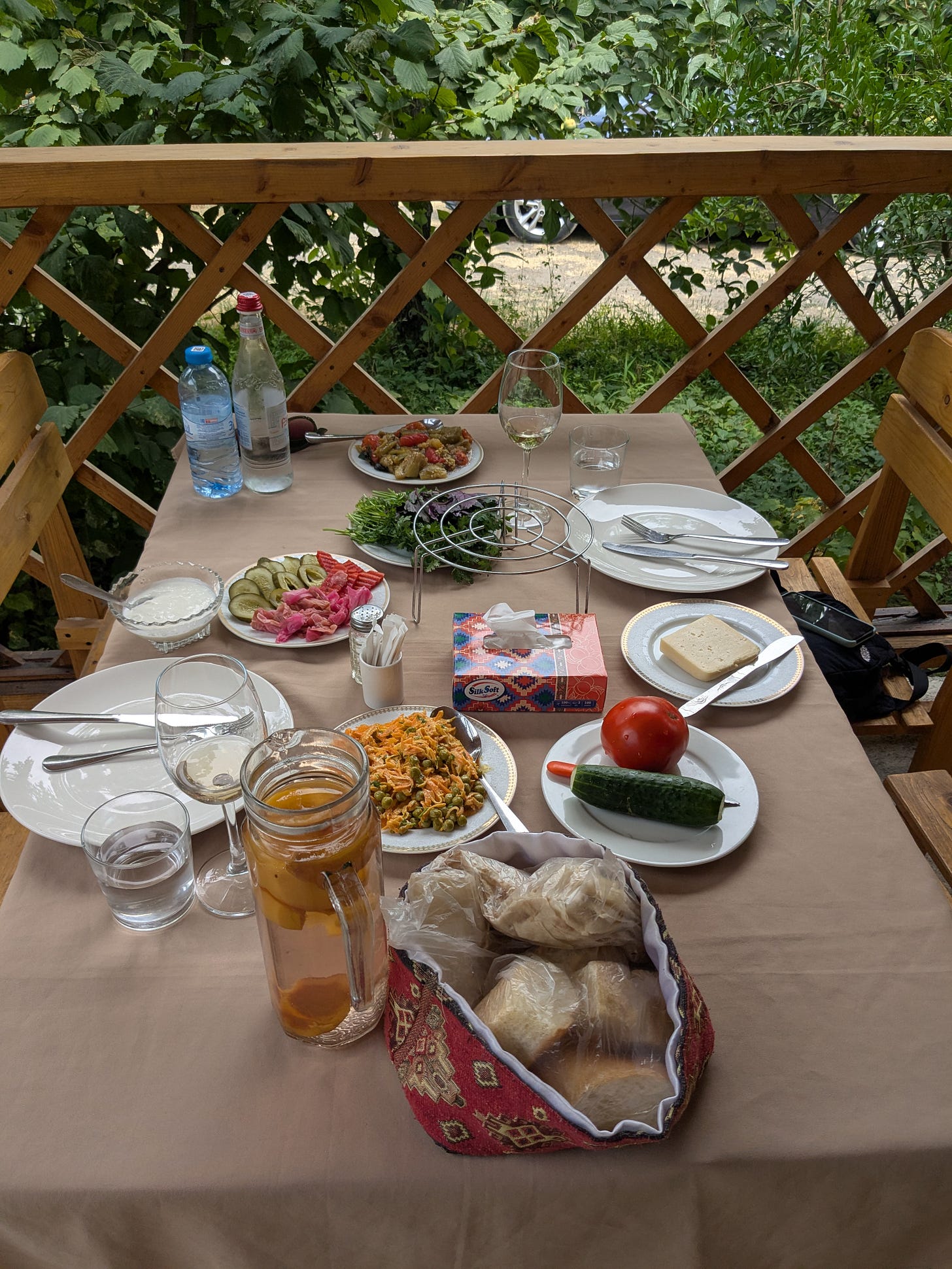 A table inside a wooden gazebo has a lunch of fresh produce displayed on a tan table cloth. There are wine glasses and white dishes set out too. 