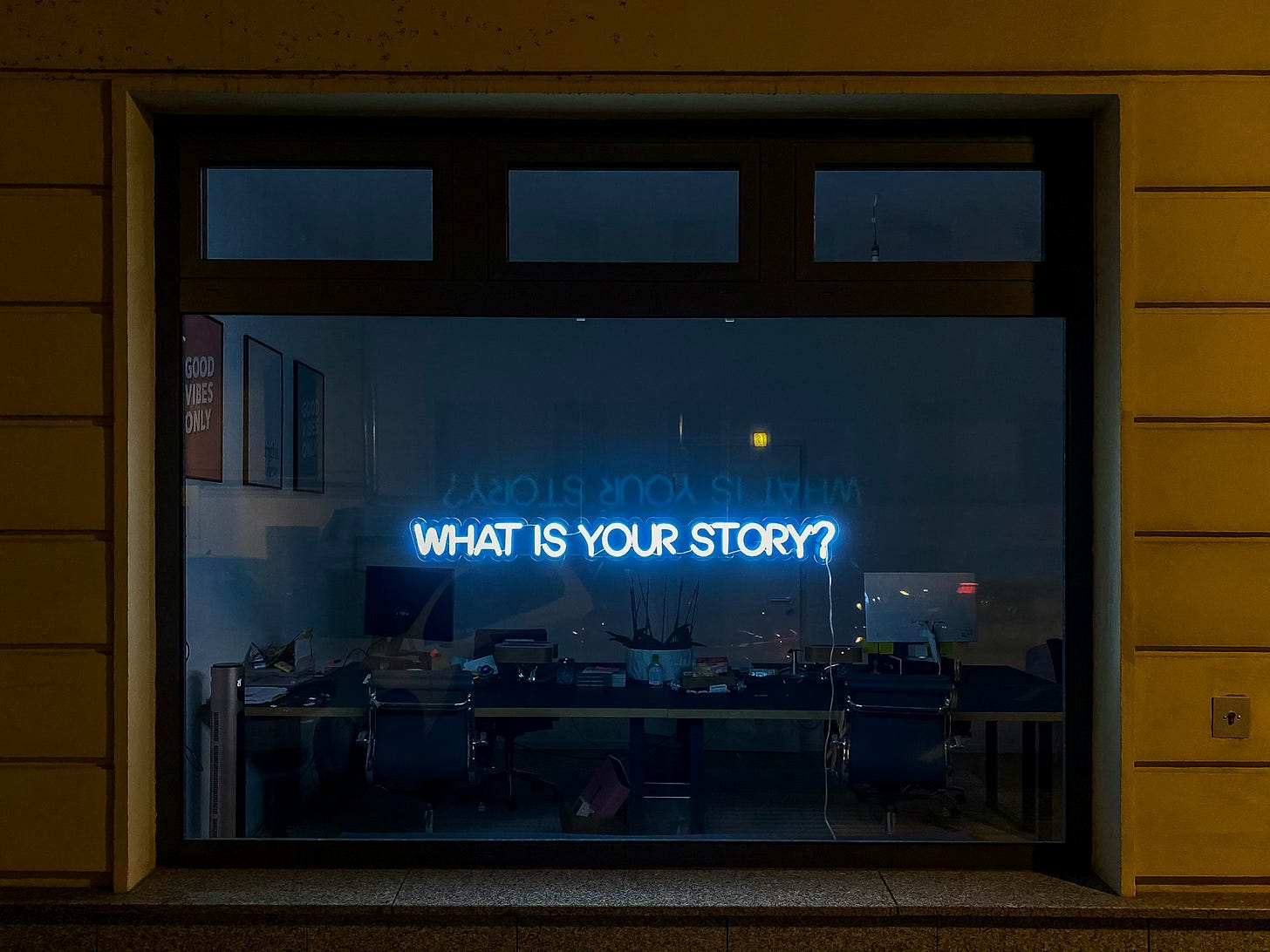 Neon text in a darkened office window reads: WHAT IS YOUR STORY?
