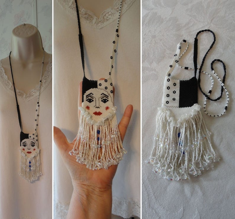 May include: A black and white beaded necklace with a fringe of white beads. The pendant is a beaded face with black and white details and a red mouth. The necklace is displayed on a white surface.
