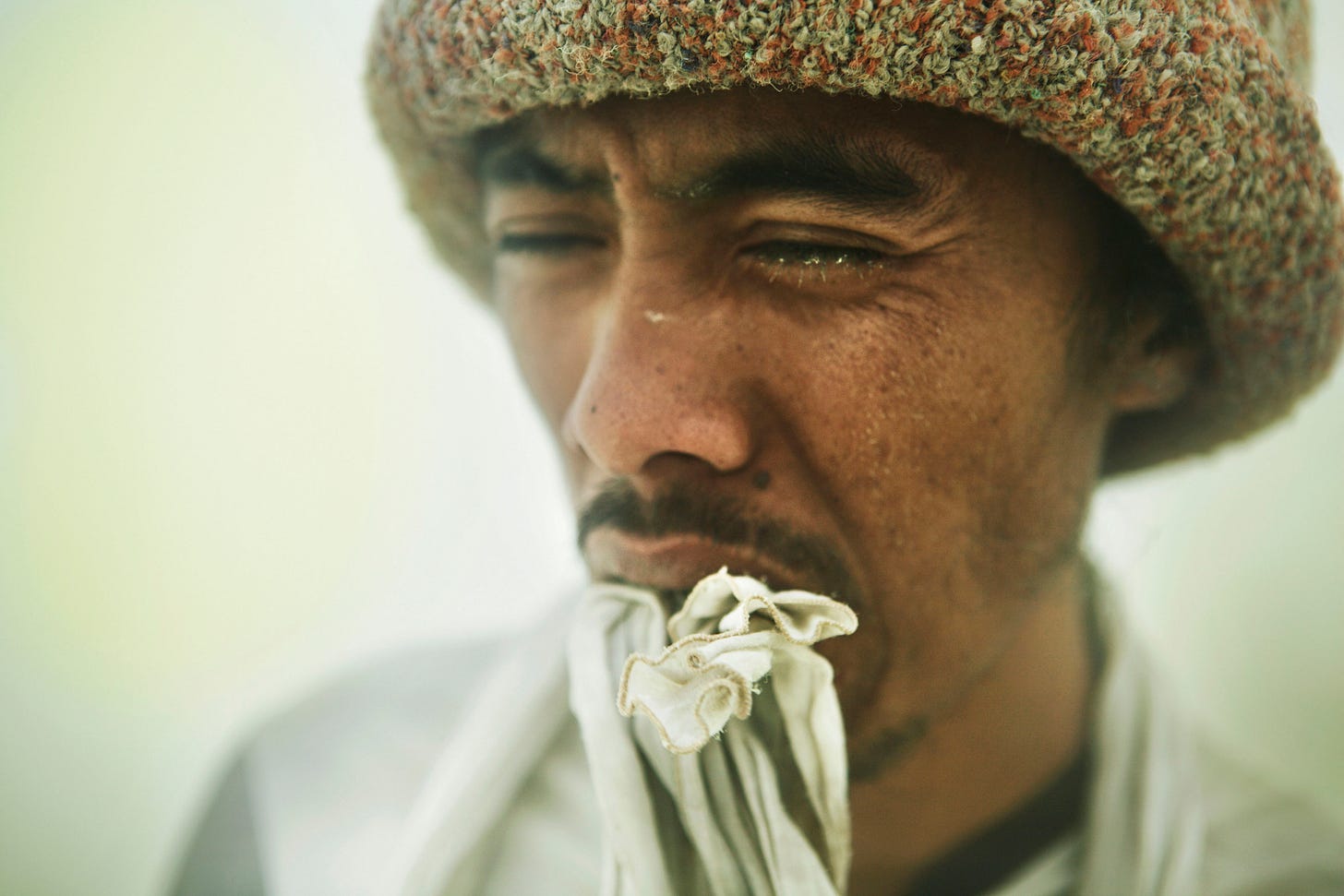A man wearing a textured, brownish hat squints intently, with a piece of patterned cloth partially covering his mouth and chin. His expression appears tense or thoughtful.