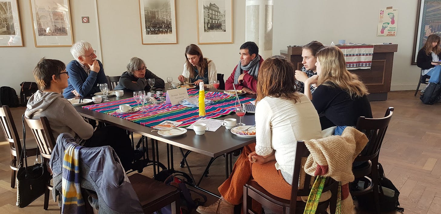David sits at a table in a large public building with 7 other people who are listening to him talk. There are plates on the table.
