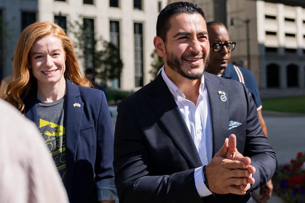 On the right, a man in a suit jacket smiles with his hands clasped. On the left, a woman in a blazer walks slightly behind him. On the right, a man in a suit jacket smiles with his hands clasped. On the left, a woman in a blazer walks slightly behind him.