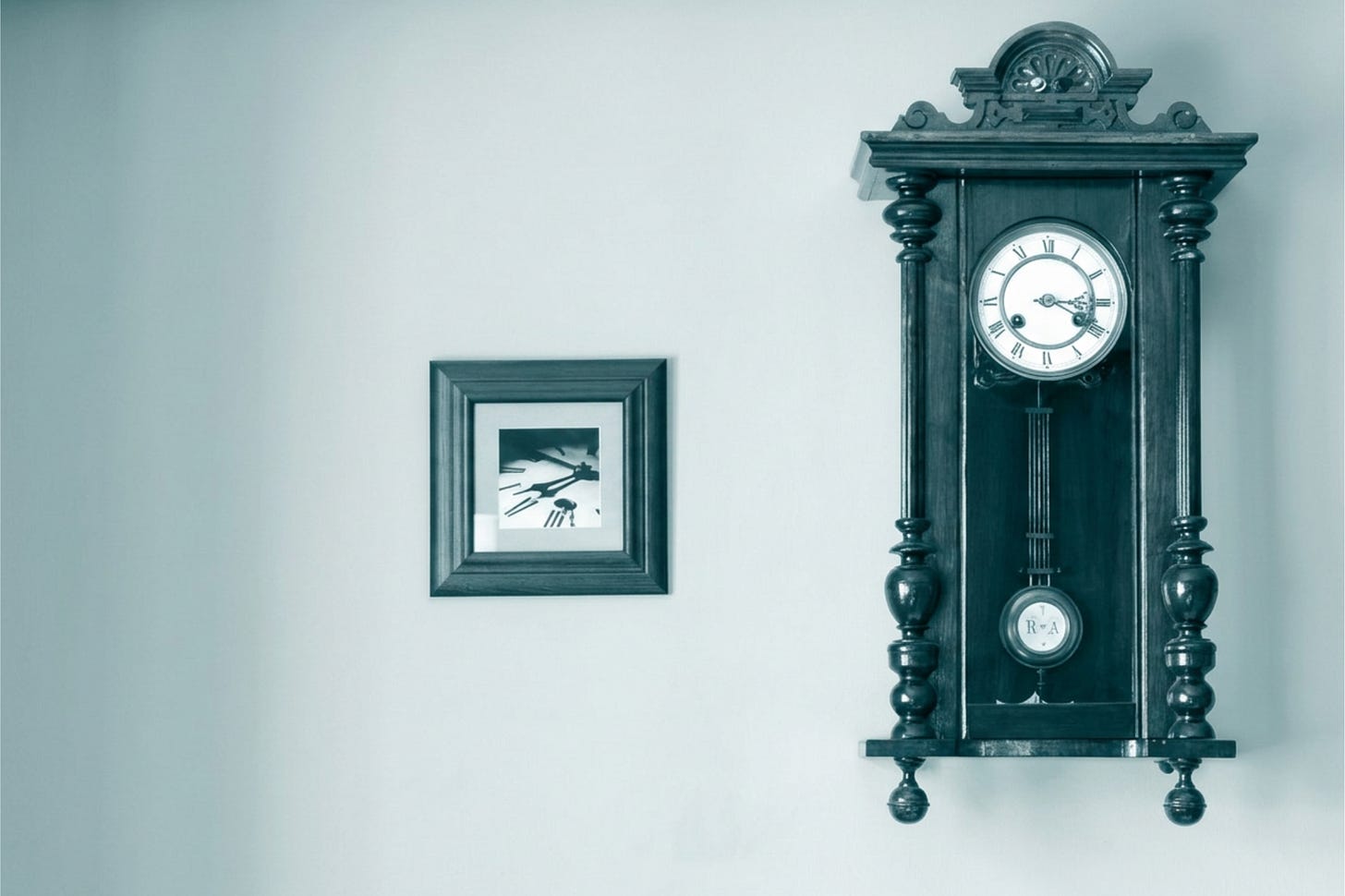 An antique wooden pendulum clock mounted on a pale wall, with a small framed picture hanging on the same wall beside it.