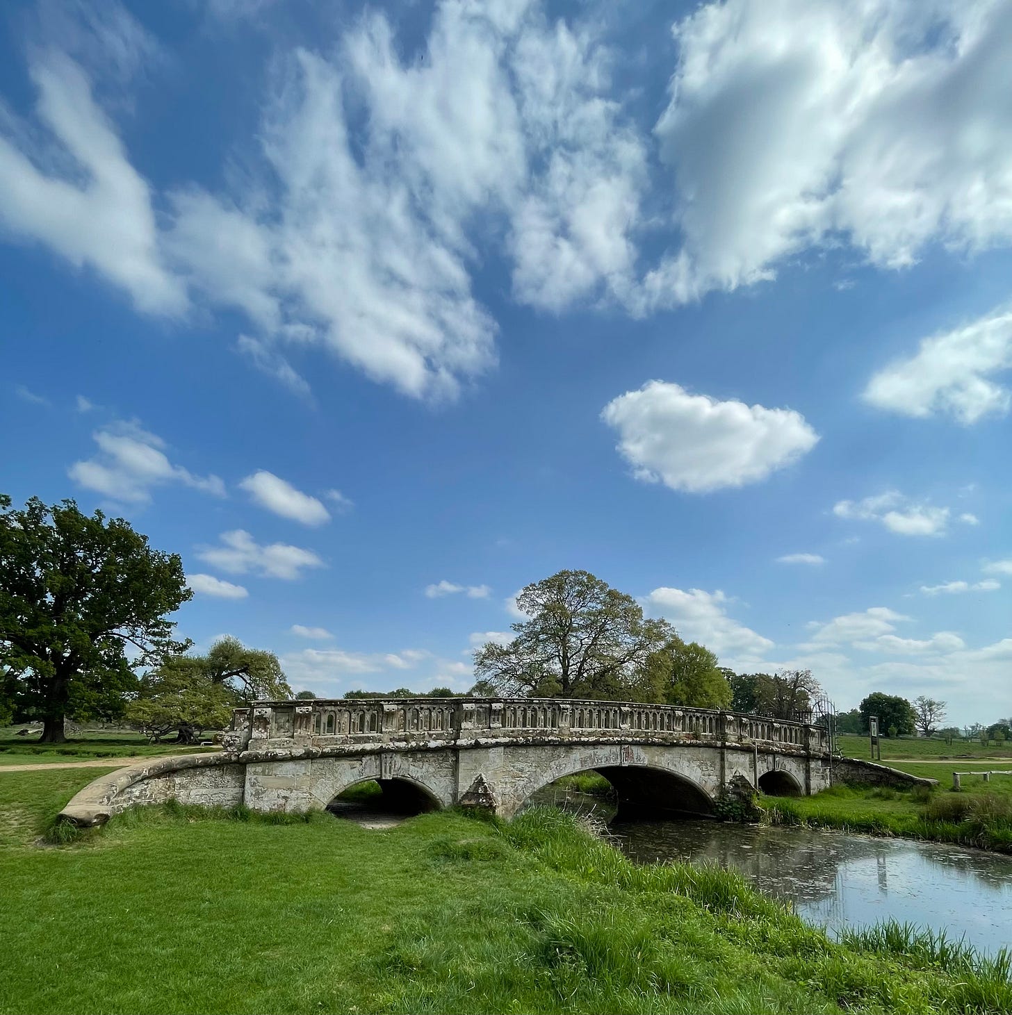 An old stone bridge over a stream, with a blue sky above and some spring-green trees