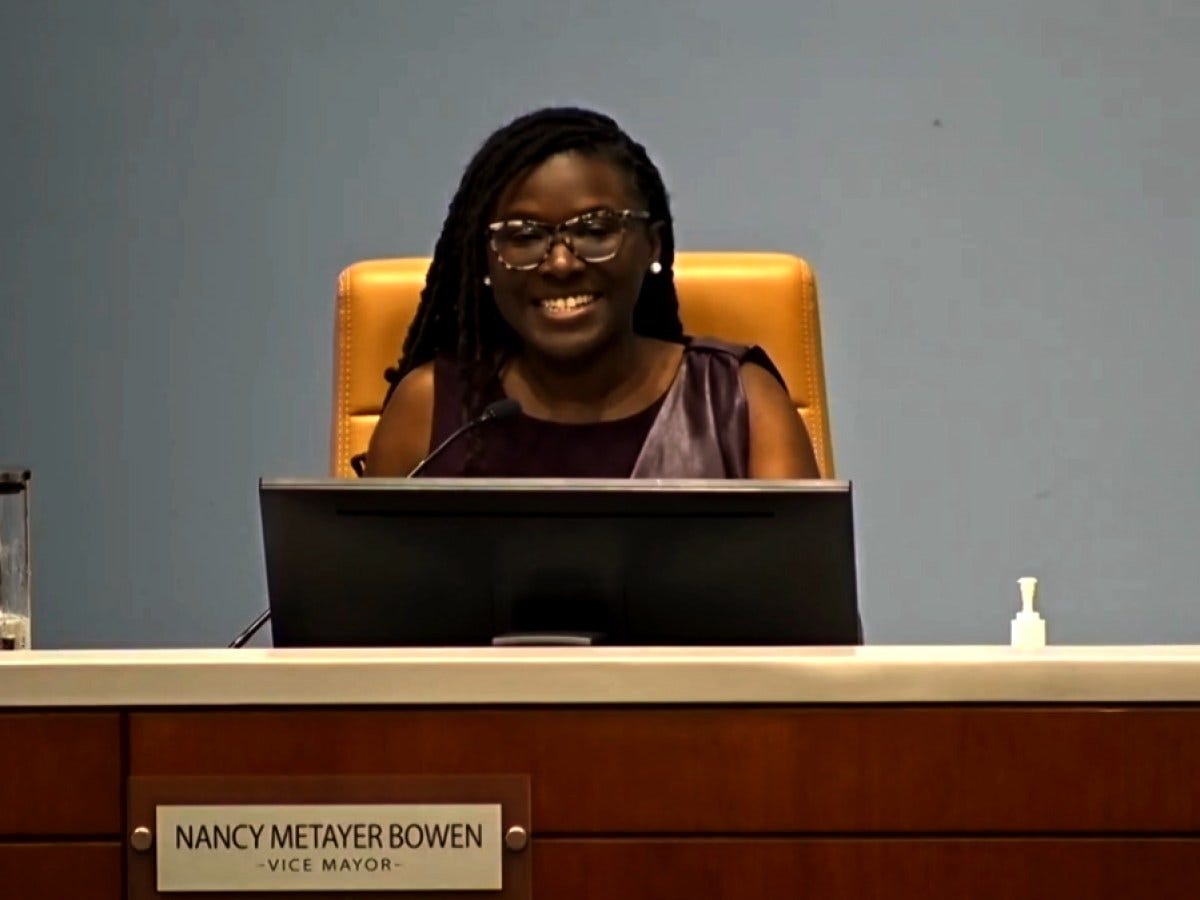 A woman wearing glasses smiles while seated behind a desk at a formal meeting or council chamber. A nameplate on the desk reads ‘Nancy Metayer Bowen – Vice Mayor.’ She sits in a tan chair behind a microphone and monitor, with a plain blue wall in the background.