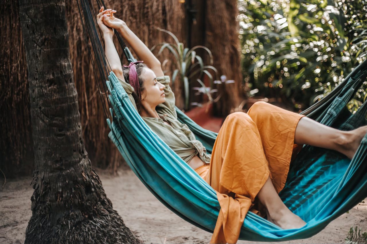 A woman in mustard color pants rest in a teal hammock near the base of a palm tree. A woman in mustard color pants rest in a teal hammock near the base of a palm tree.