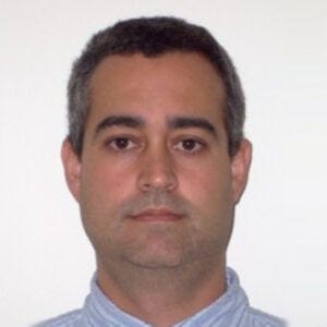 Homem de aparência adulta, com cabelo curto e escuro levemente grisalho nas laterais, pele clara, sobrancelhas marcadas e expressão neutra.