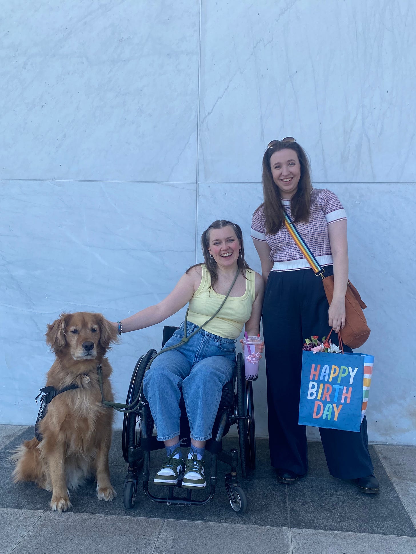 meredith, a wheelchair user wearing a yellow top, light wash jeans, dunks and a hands free light green leash, scout a golden retriever, and lily wearing a knit short sleeve top and navy oants smile infront of a marble wall. 