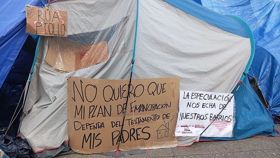 Tienda de campaña de acampada reivindicativa en una plaza pública, adornada con carteles manuscritos de protesta sobre vivienda y especulación. Tienda de campaña de acampada reivindicativa en una plaza pública, adornada con carteles manuscritos de protesta sobre vivienda y especulación.