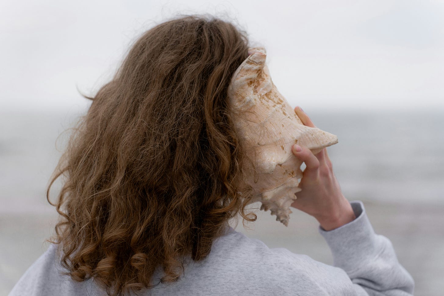 A person with long, wavy brown hair holds a large conch shell to their ear while standing by the sea on a cloudy day, symbolizing quiet listening and connection with inner sound.