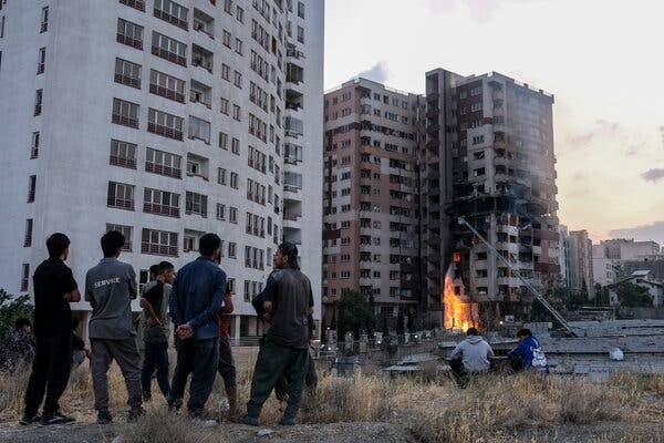 A group of people watch a burning apartment building from a distance.