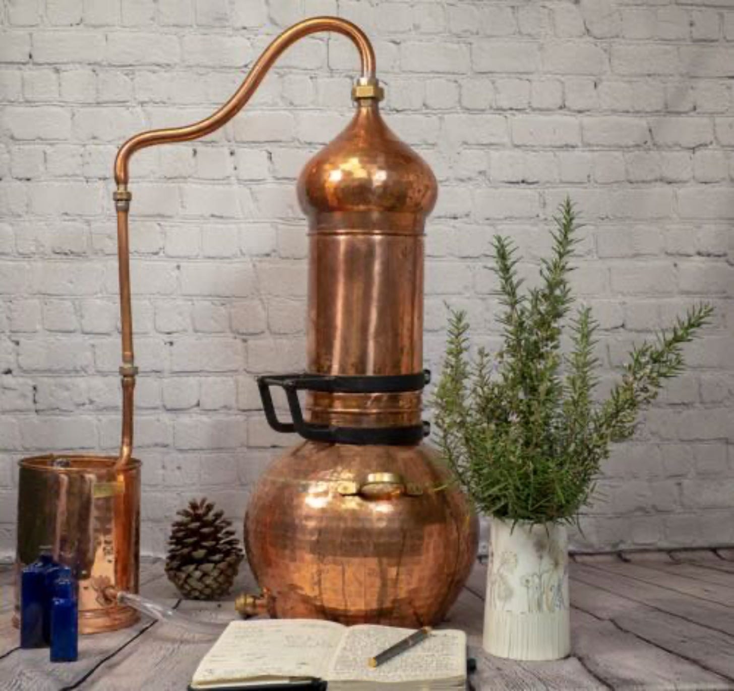 A copper still stands next to bunches of rosemary and a recipe book