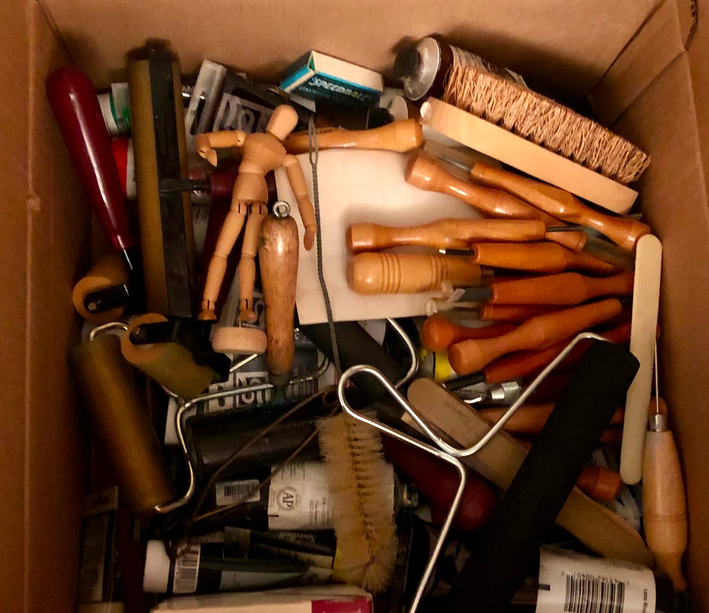 A box of loose art supplies, mostly print-making tools, and in one corner is a face-down jointed wooden mannequin.