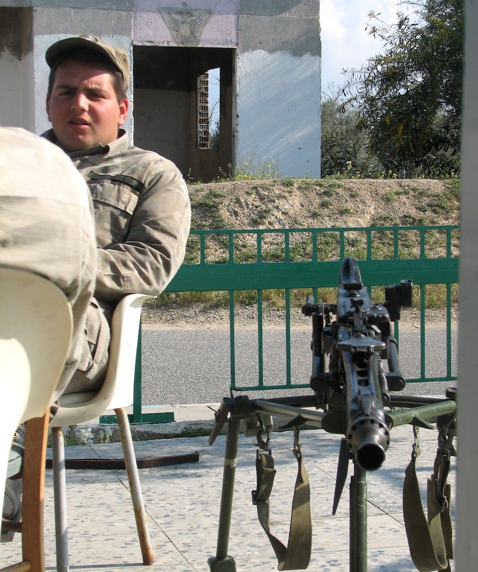 a guy in the army sitting in a chair chatting next a machine gun set up on the ground