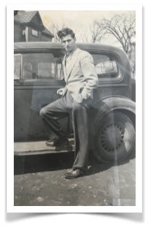 Black and white photo of a dark-haired man wearing a suit coat and tie, posing next to a car.