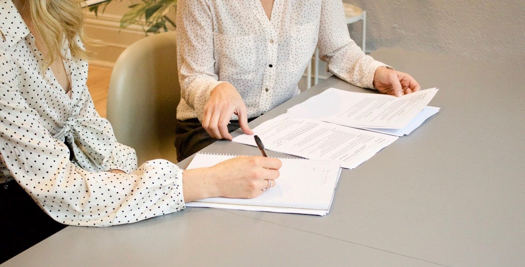 woman signing on white printer paper beside woman about to touch the documents woman signing on white printer paper beside woman about to touch the documents