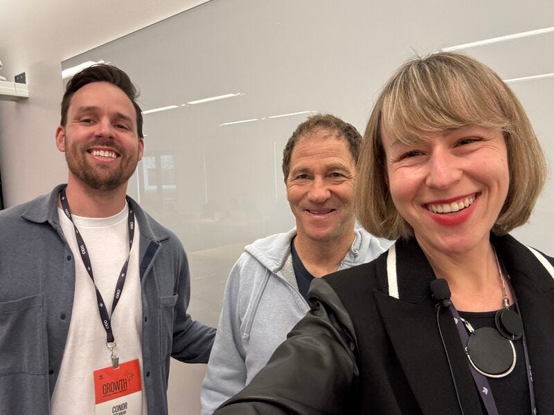 Three people smiling at the camera in an indoor conference or office setting, taking a casual group selfie. Two men stand on the left and center wearing lanyards and casual layers; a woman on the right with a short bob haircut wears a black jacket and red lipstick. A glass wall and bright, modern interior are visible in the background.