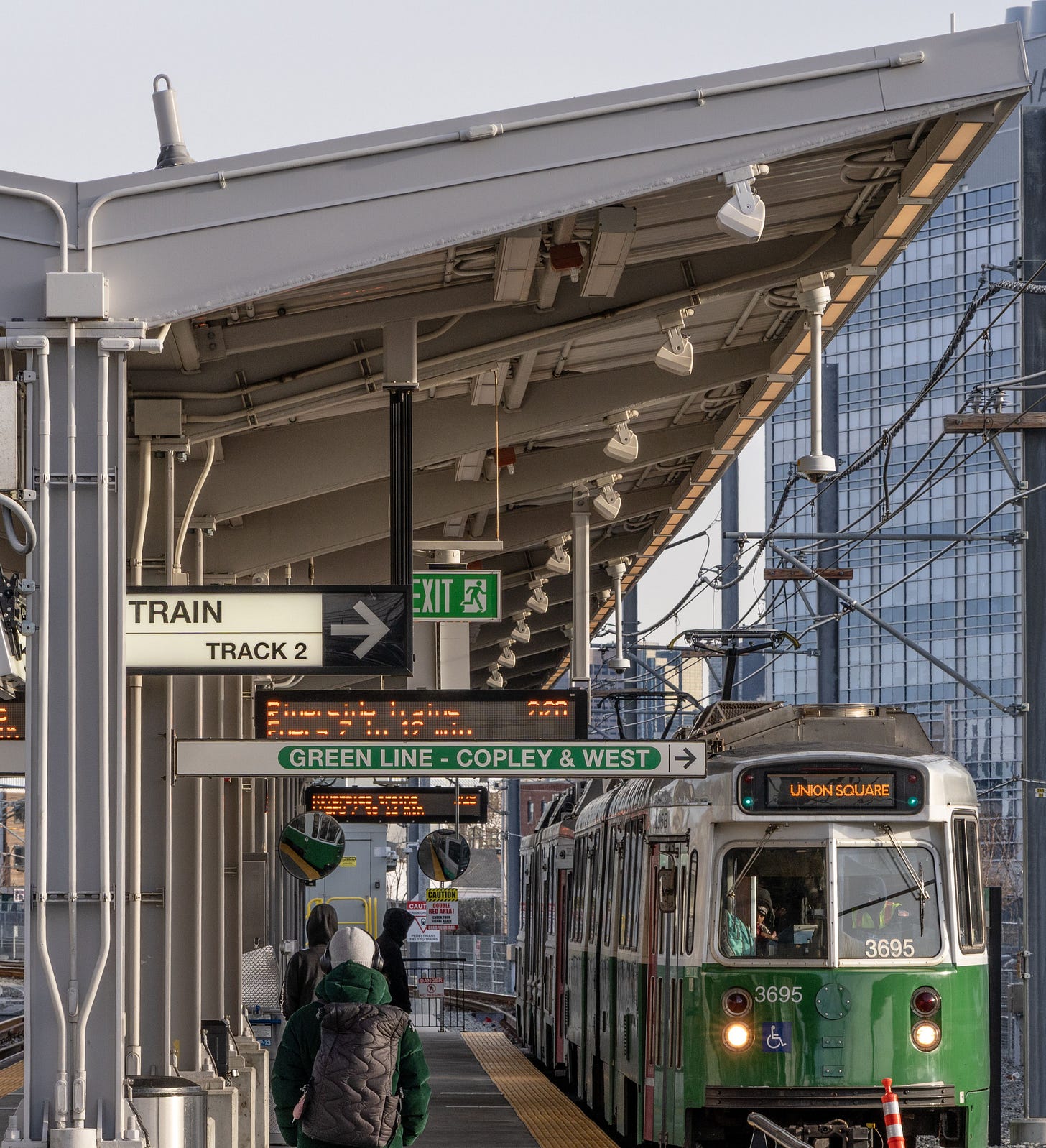 Green Line train at a Somerville station platform with signs and passengers. Green Line train at a Somerville station platform with signs and passengers.