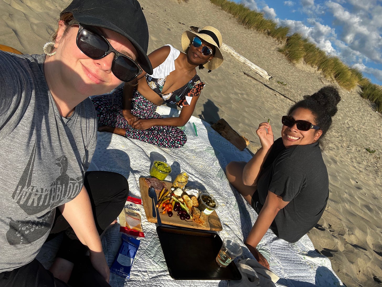 Amber, Nafissa, and Jackie sit on a blanket on the beach with a charcuterie board spread in front of them. Amber takes a group selfie of the three of them.