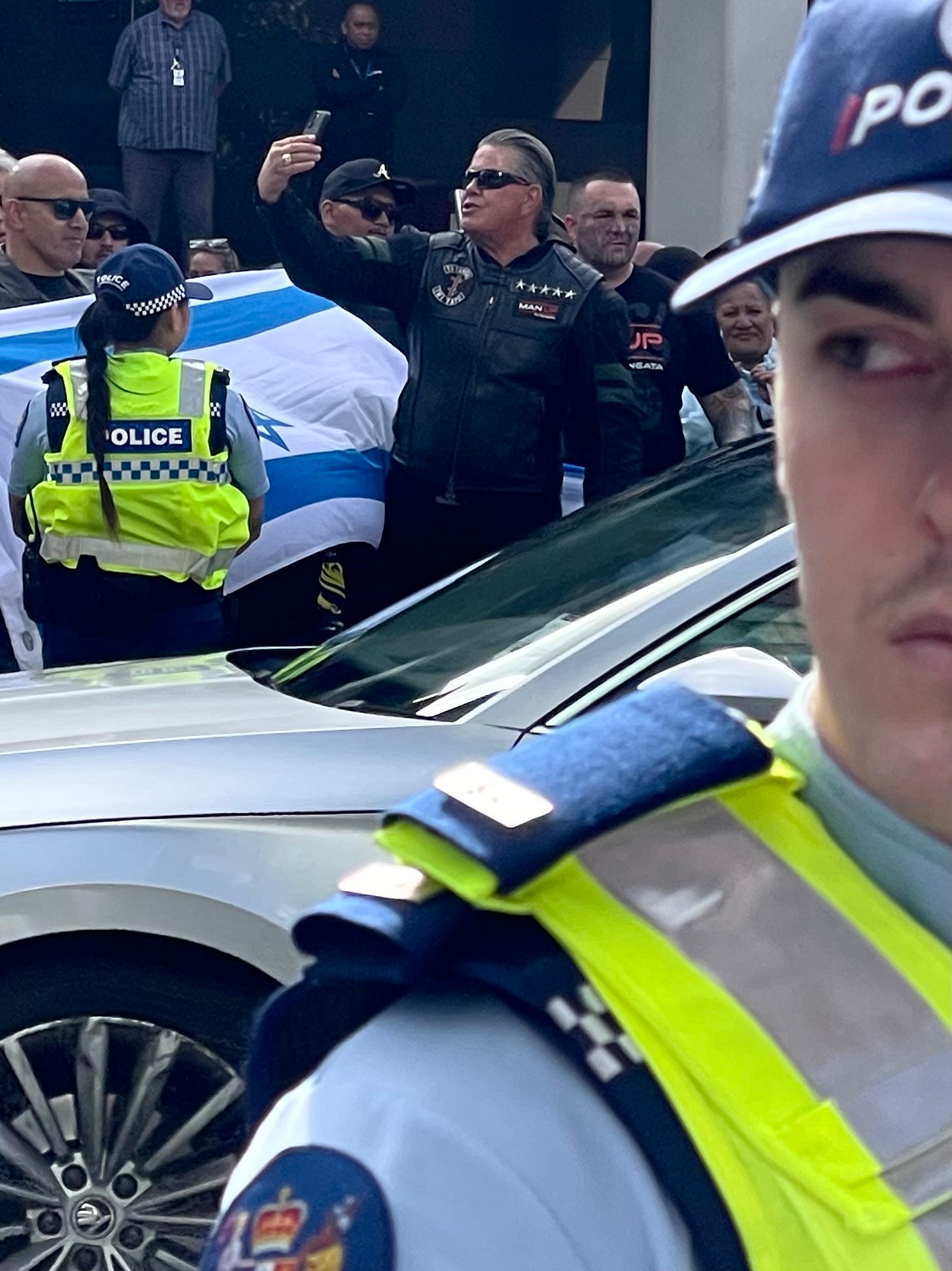 A crowded street scene where police officers in high-visibility vests stand in the foreground and midground, partially blocking the view. Behind them, a group of people gathers closely together; one man wearing a black leather vest and sunglasses holds up a smartphone as if recording or taking a photo. An Israeli flag is visible among the crowd. A police car is parked between the officers and the group, and additional onlookers stand in the background near a building. A crowded street scene where police officers in high-visibility vests stand in the foreground and midground, partially blocking the view. Behind them, a group of people gathers closely together; one man wearing a black leather vest and sunglasses holds up a smartphone as if recording or taking a photo. An Israeli flag is visible among the crowd. A police car is parked between the officers and the group, and additional onlookers stand in the background near a building.