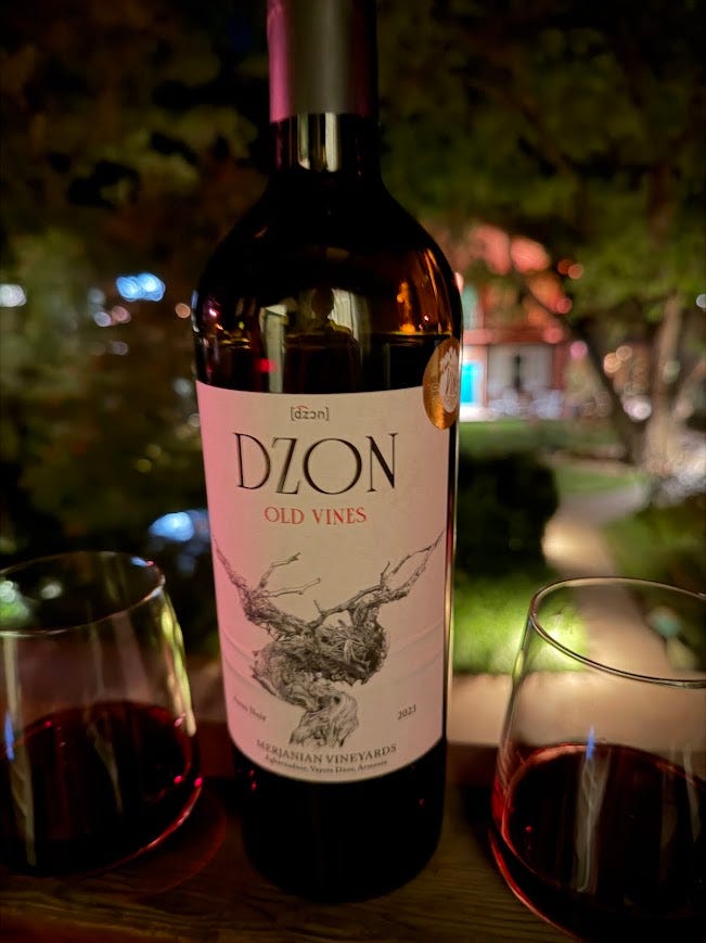 A bottle of Dzon Old Vines Areni Noir from Merjanian Vineyards on an outdoor table at night, flanked by two glasses of red wine, with string-lit trees in the background.