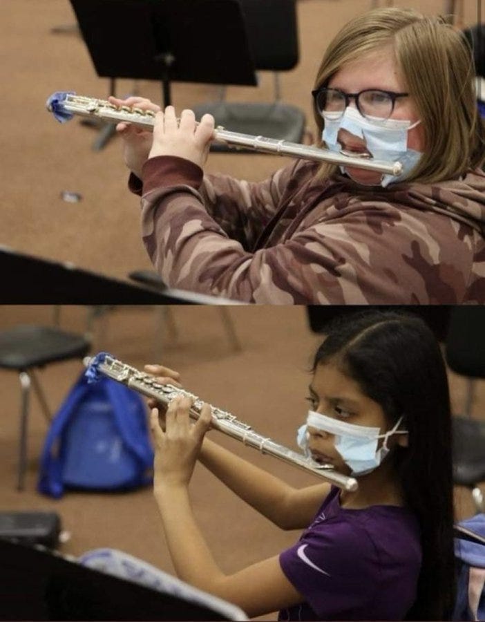 Two individuals playing flutes in a room with music stands and chairs. Both are wearing camouflage jackets and face masks, holding silver flutes with blue ends. One person has long brown hair and glasses, the other has long black hair and a purple shirt. Two individuals playing flutes in a room with music stands and chairs. Both are wearing camouflage jackets and face masks, holding silver flutes with blue ends. One person has long brown hair and glasses, the other has long black hair and a purple shirt.