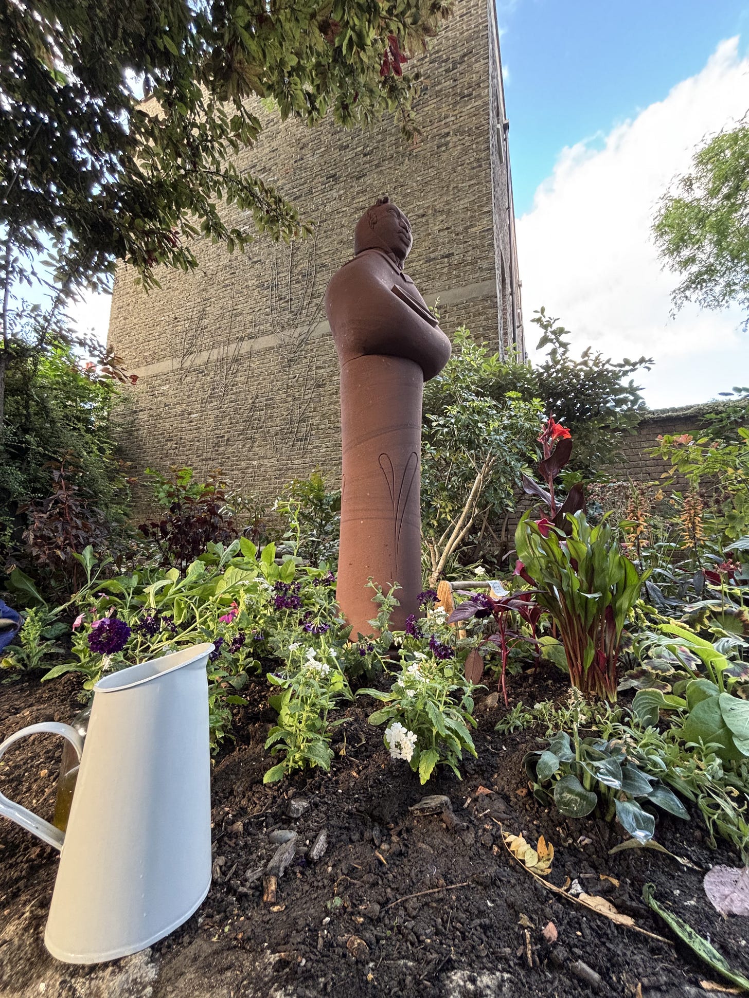 A tall clay statue of a Black woman healer nestles in a healing garden of plants, sheltered by tall trees and the wall of a tall brick building. In the foreground a white enamel water jug sits on the soil