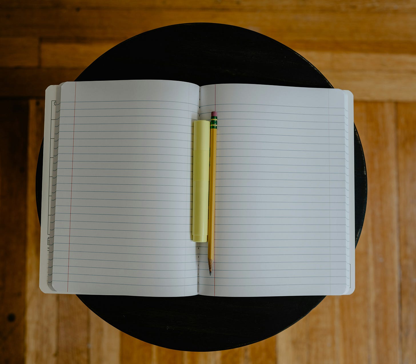 A paper notebook lays open and flat on a small, circular black table. A pencil and highlighter sit in the middle of the book, holding open the pages. A paper notebook lays open and flat on a small, circular black table. A pencil and highlighter sit in the middle of the book, holding open the pages.