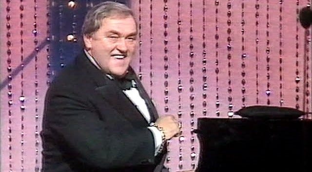 Les Dawson at at a piano, turning to camera and smiling while ‘selling’ a gag