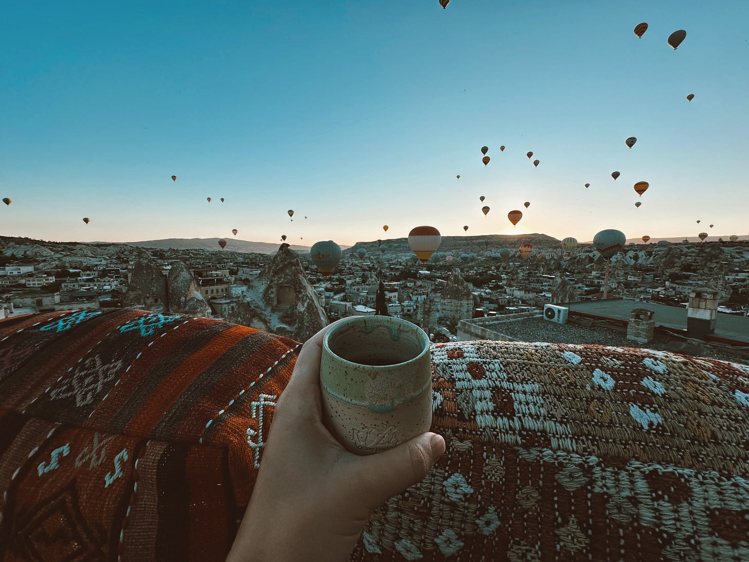A hand holding a ceramic mug overlooking a scenic view of Cappadocia, Turkey at sunrise. Dozens of hot air balloons float over a landscape of ancient rock formations and rooftops covered in patterned Turkish rugs. Symbolizes the freedom and beauty of a location-independent writing life.
