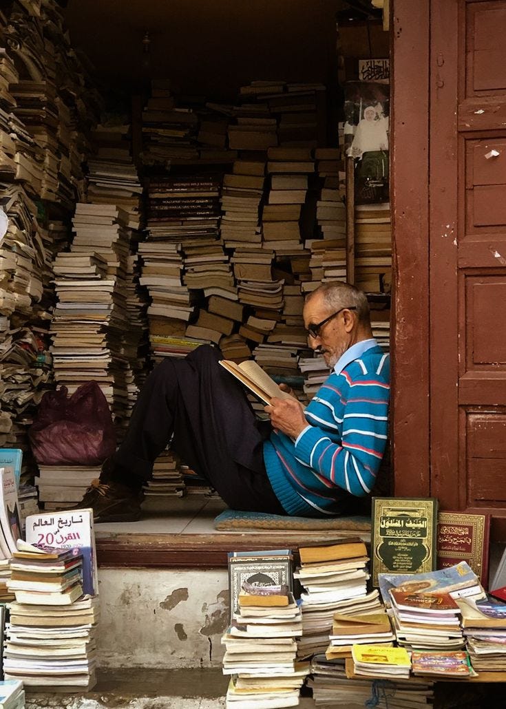 This may contain: a man sitting on the ground reading a book in front of a pile of books This may contain: a man sitting on the ground reading a book in front of a pile of books