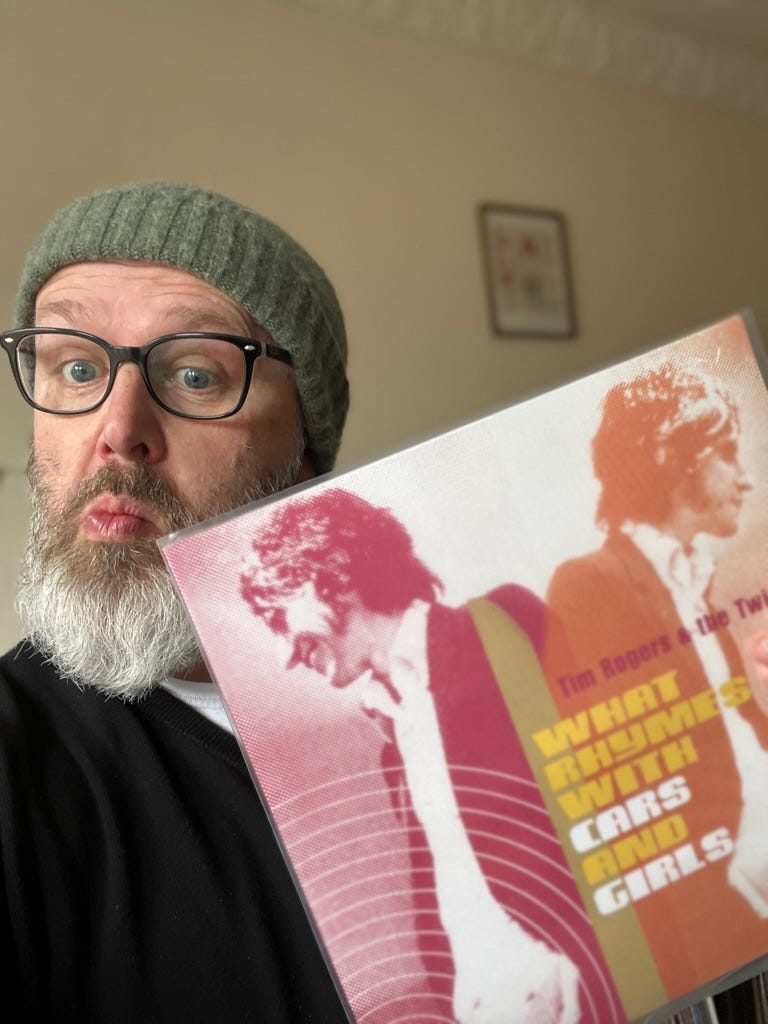 Tom wearing a beanie and holding a Tin Roger & the Twin Set album call What Rhymes With Cars And Girls