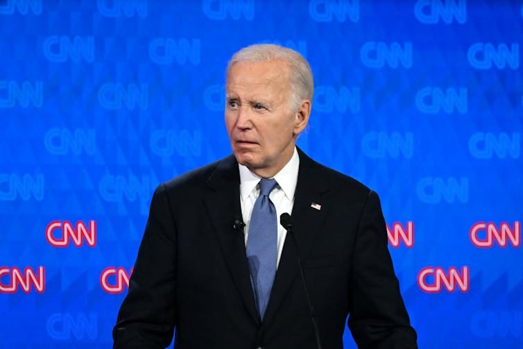 A gray-haired man in a dark suit, standing on a stage, looks at something to his right. A gray-haired man in a dark suit, standing on a stage, looks at something to his right.