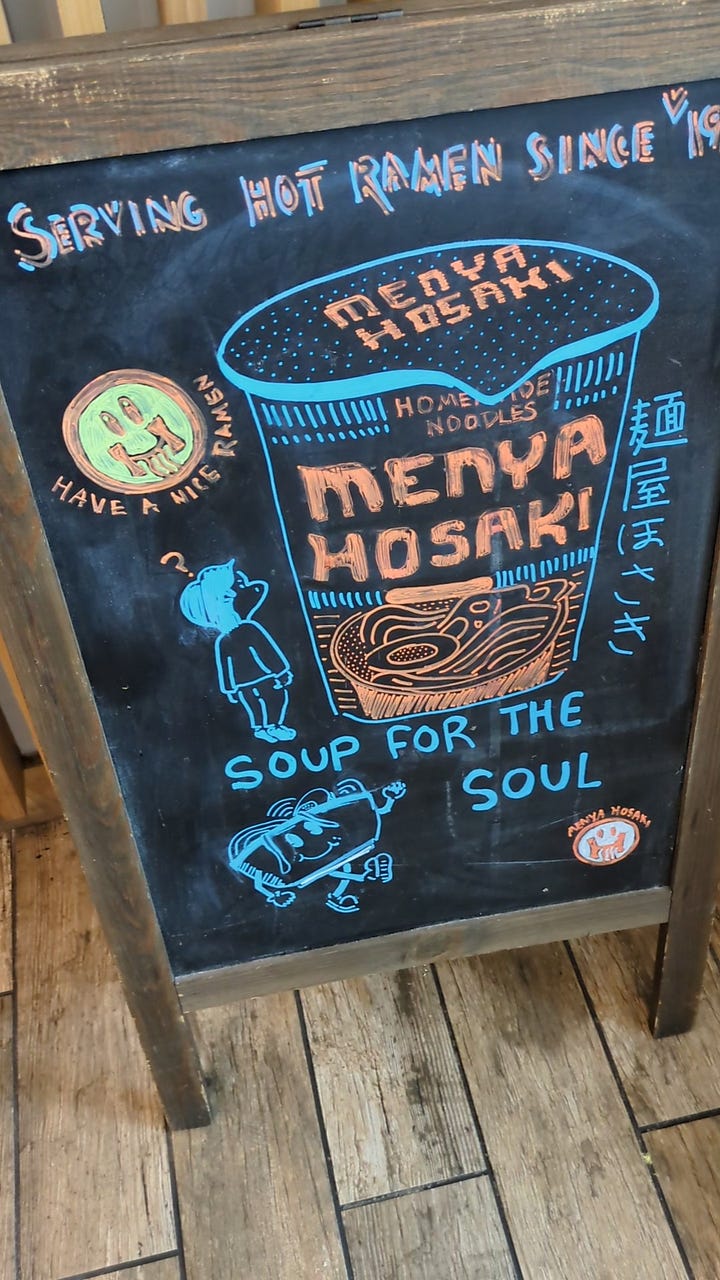 Art in menya hosaki, a take on the famous 'Great Wave' painting of Mount Fuji with Dogs surfing, and a hand-drawn chalkboard sidewalk sign with menya hosaki written on something that looks like a cup of noodles and little faces and characters.