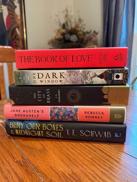 A stack of books on a wooden table, including: The Book of Love by Kelly Link, One Dark Window by Rachel Gillig, The City of Brass by S.A. Chakraborty, Jane Austen's Bookshelf by Rebecca Romney, and Bury Our Bones in the Midnight Soil by V.E. Schwab. 
