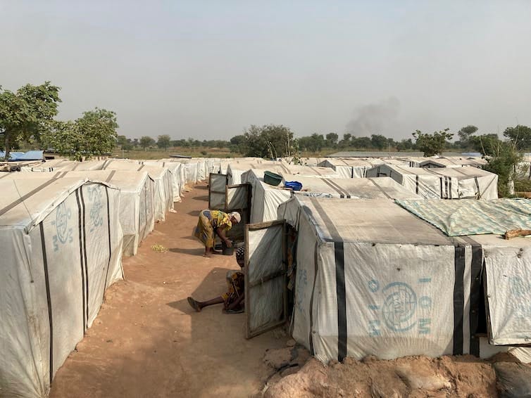 People sit outside square white tents in the middle of a dirt-packed field. People sit outside square white tents in the middle of a dirt-packed field.