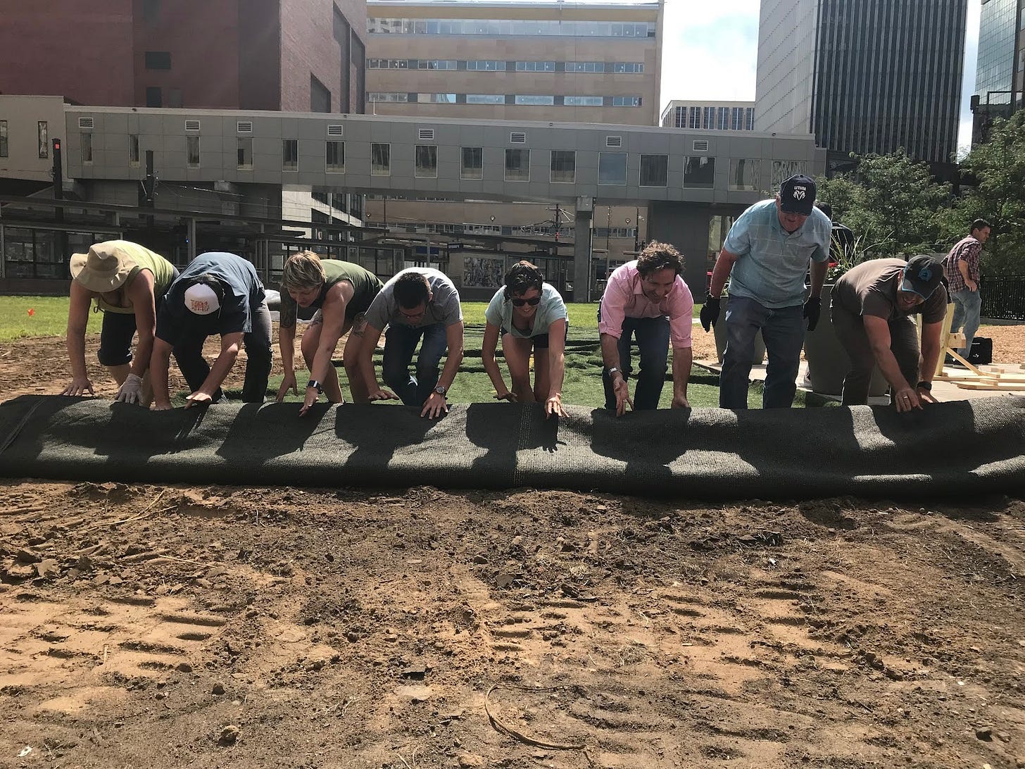 A line of volunteer workers roll out a length of astroturf with an empty lot and downtown buildings in the background.