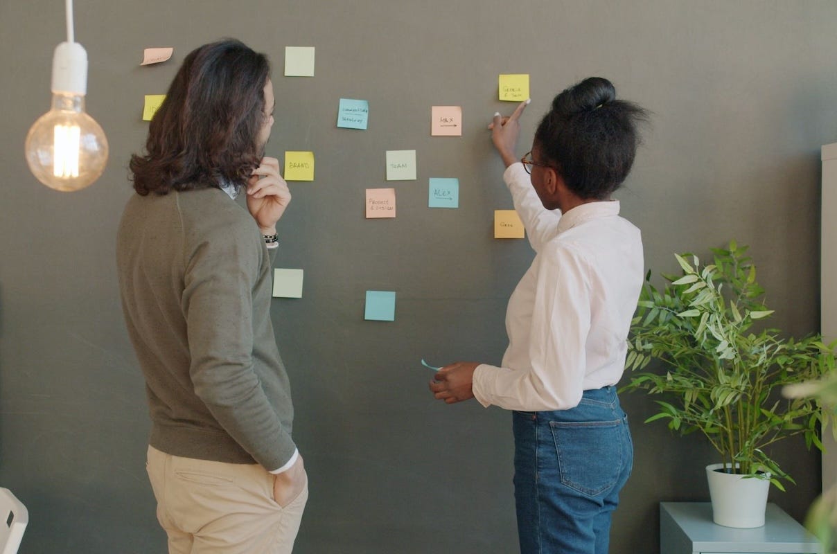 Two marketing professionals collaborating and brainstorming a B2B messaging framework using colorful sticky notes on a wall. Two marketing professionals collaborating and brainstorming a B2B messaging framework using colorful sticky notes on a wall.