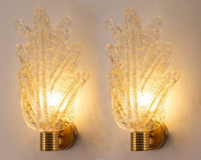 May include: Two gold wall sconces with clear glass shades. The glass shades are textured and have a flame-like shape. The sconces are lit and the light is shining through the glass.