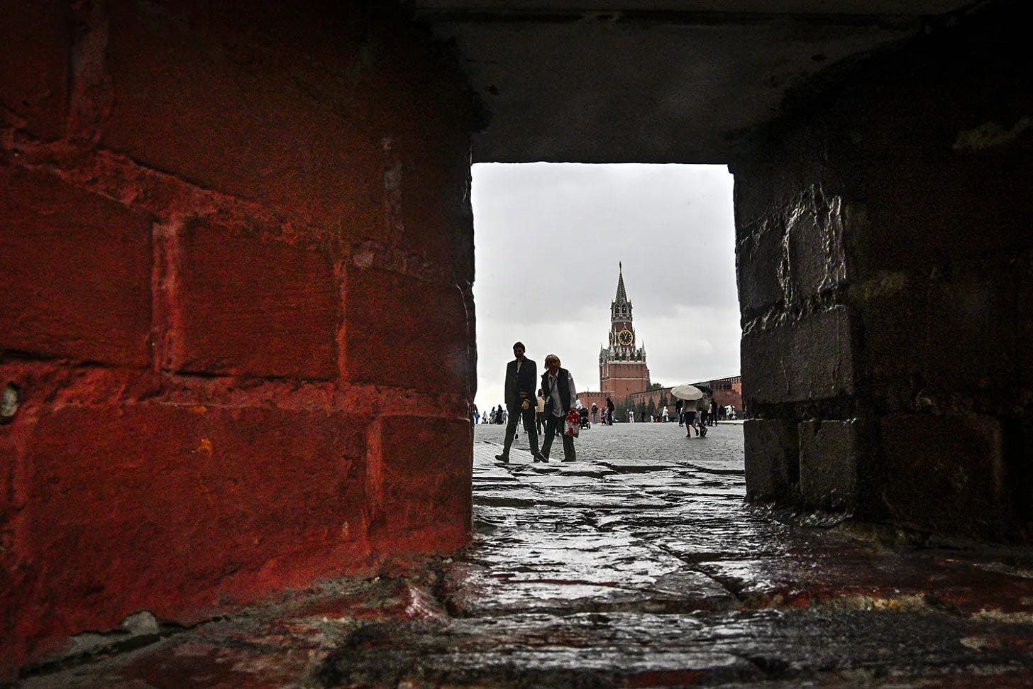 A view framed by red brick structure in the foreground, showing two people walking across a large open plaza toward a high tower topped with a clock in the distance under a cloudy sky. A view framed by red brick structure in the foreground, showing two people walking across a large open plaza toward a high tower topped with a clock in the distance under a cloudy sky.