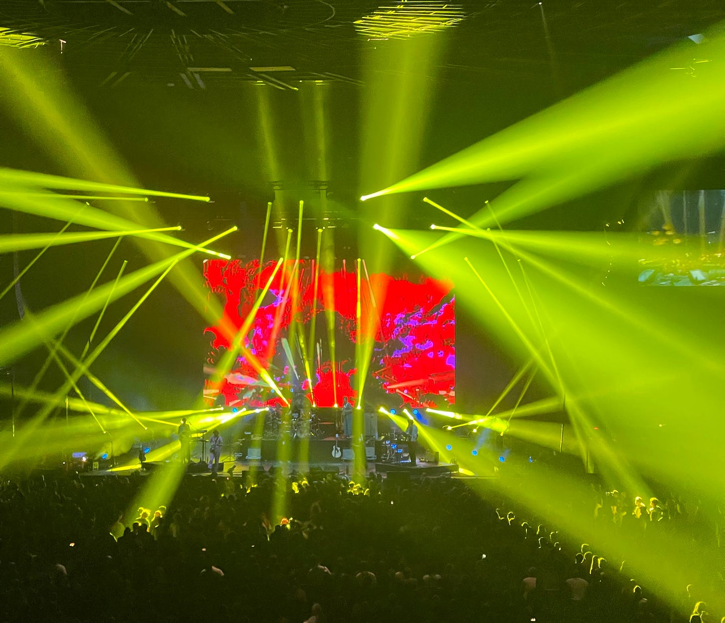 Wide shot of the stadium in the middle of the King Gizzard & the Lizard Wizard show. Laser lights shoot in every direction and multicolored psychedelic image effects flash across a large screen behind the band.