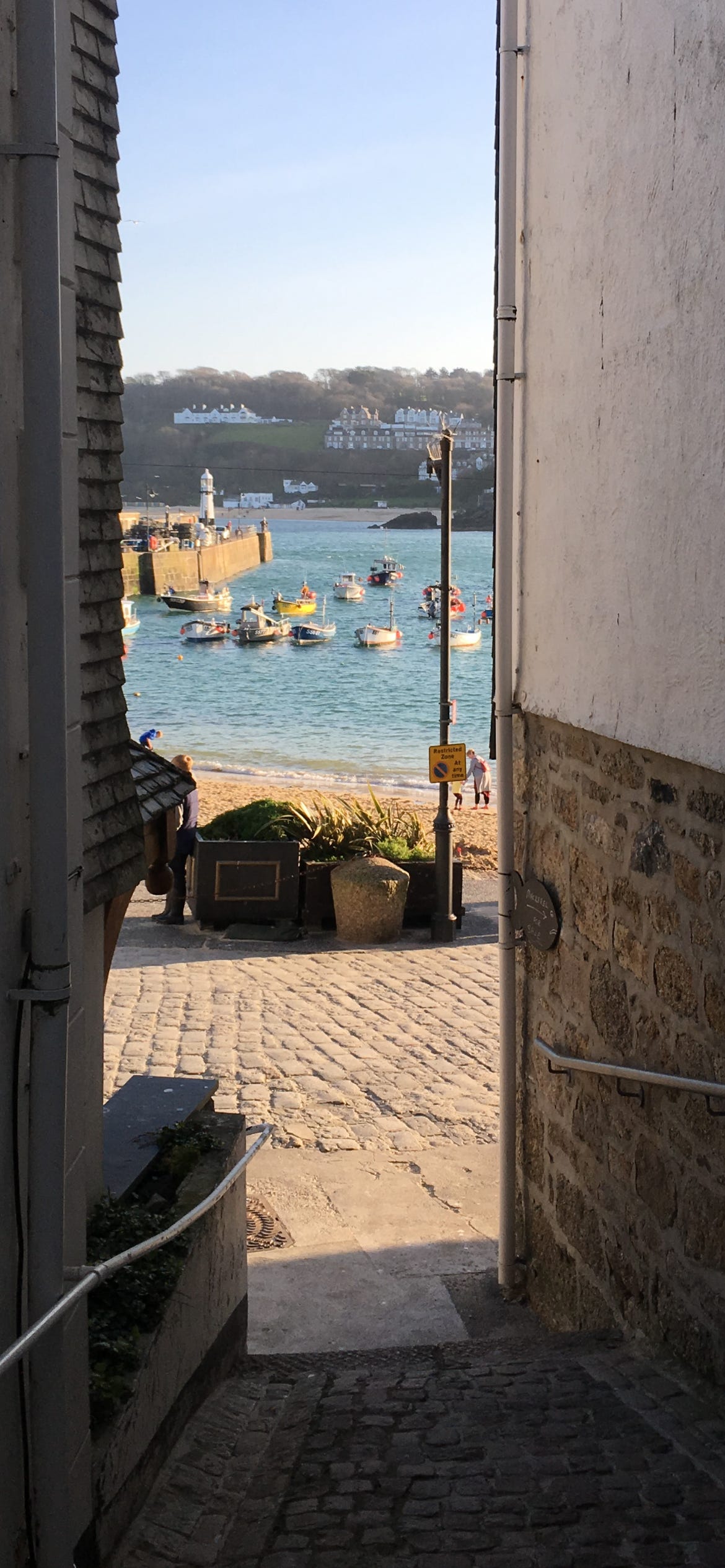 A view to St Ives harbour down a narrow alleyway between two buildings. The sun is shining in the harbour and the sky is a bright summer blue. The view beckons us towards it.